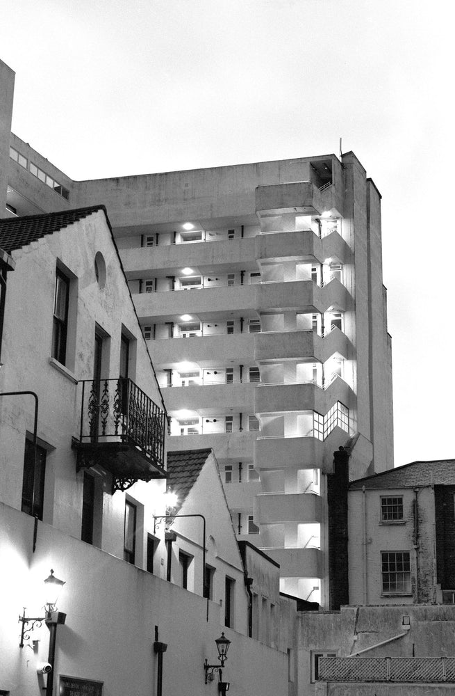 A white apartment block with stacked lit balconies rising behind older rooftops at dusk, black and white photograph shot on the Intrepid Super 120