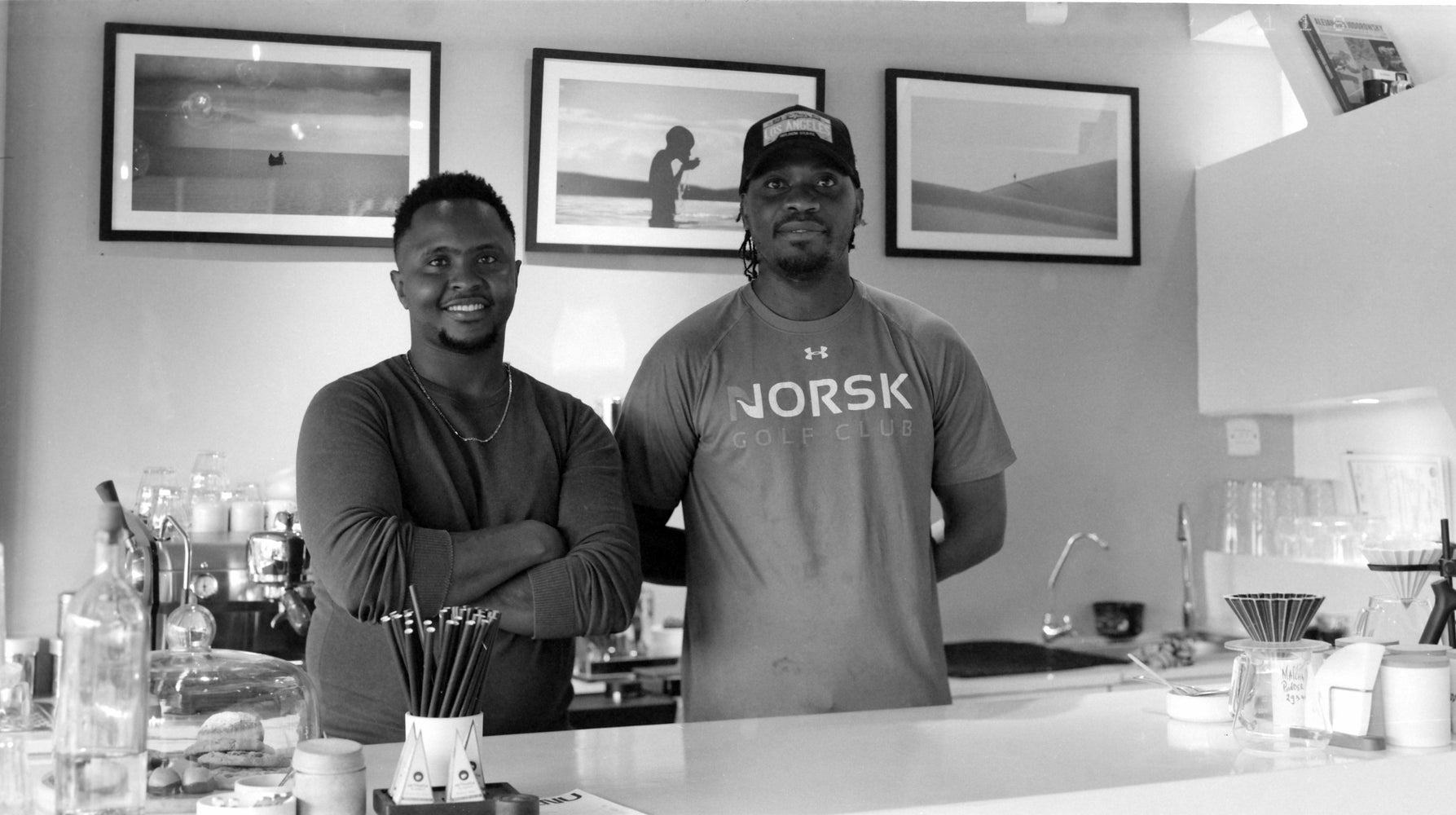 Two men standing behind a coffee bar counter with framed photographs on the wall, black and white photograph shot on the Intrepid Super 120