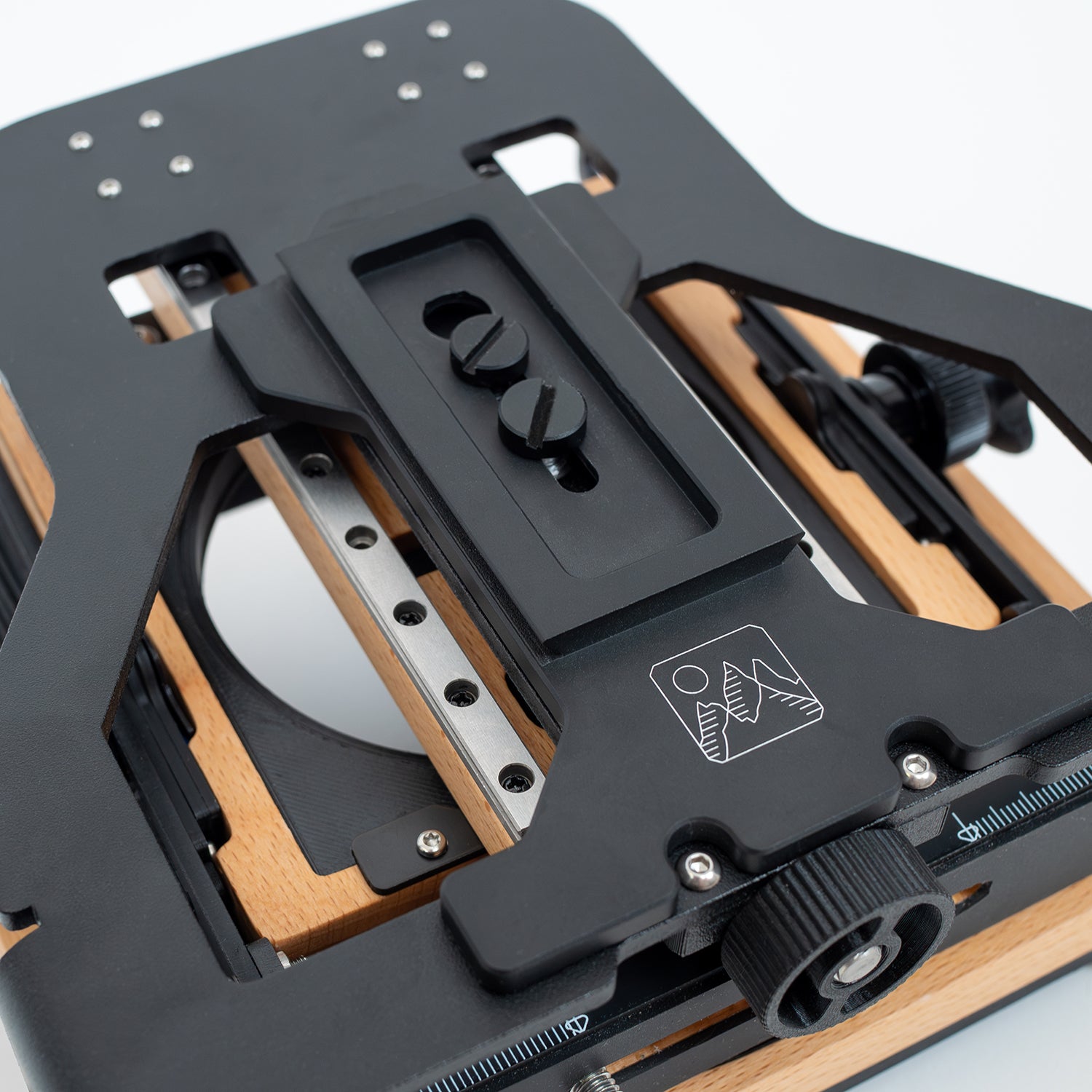 A black Intrepid Arca Tripod Plate (mounted on an Intrepid 4x5 camera) made of aluminium with a 1/4-20 screw and a 3/8-16 screw, designed to hold and secure cameras on an Arca style tripod head.