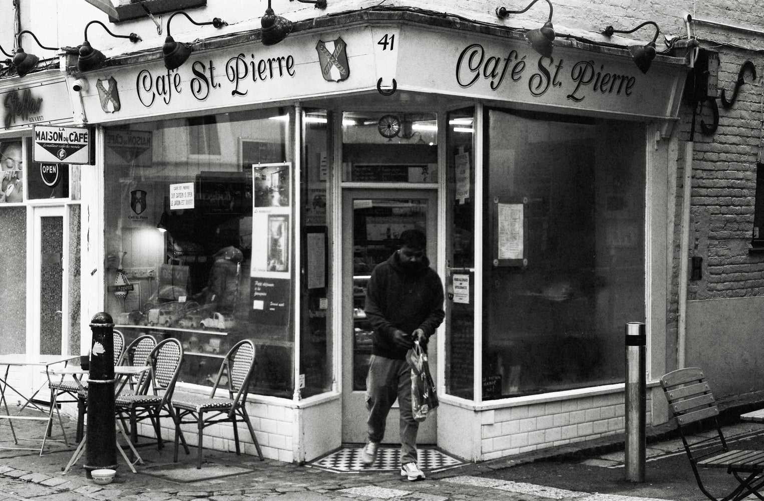 A man leaving Café St. Pierre, a corner café, carrying a shopping bag, black and white photograph shot on the Intrepid Super 120